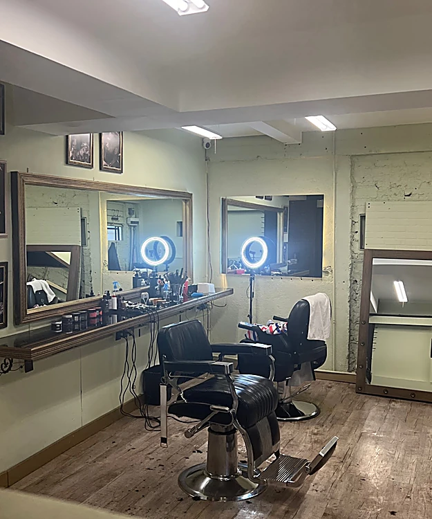 Sala de muros verde salvia con dos sillas de barbero negras, espejos y aros de luz.