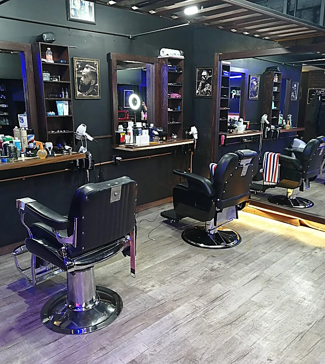 Interior con muros negros, sillones de barbero cromados, espejos y repisas con productos.