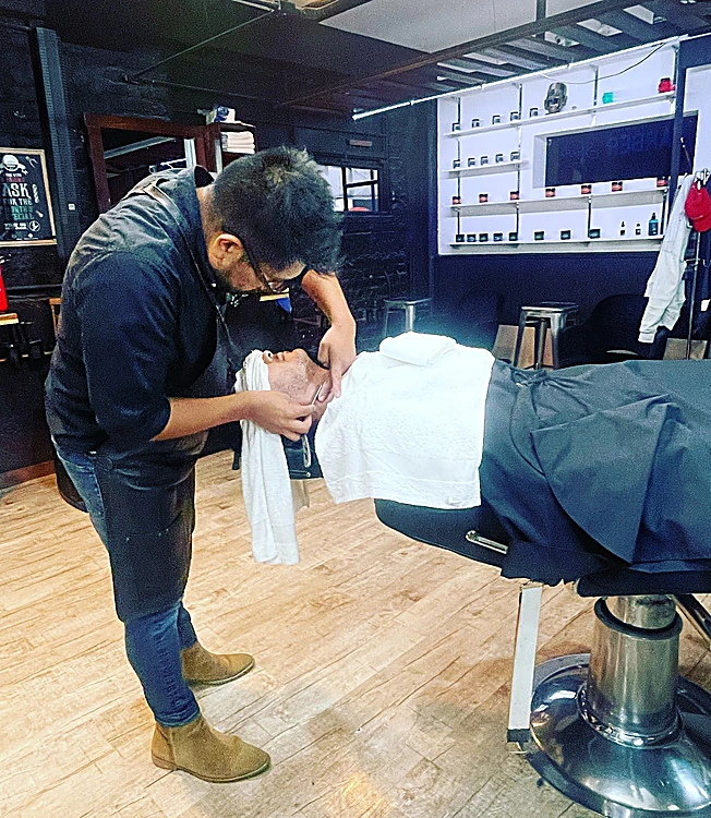 Barbero preparando un afeitado con toalla caliente; el cliente está recostado y cubierto por la toalla blanca.